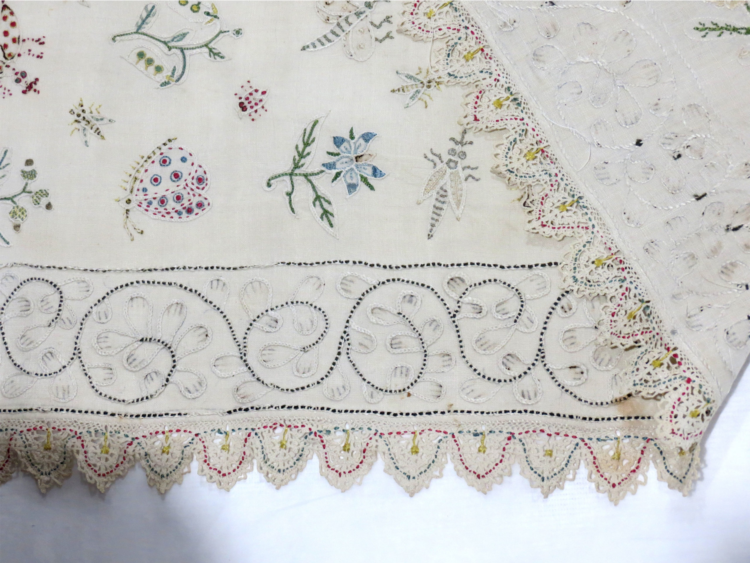 White linen embroidered in coloured silks and white linen threads with bands of birds, insects, flowers, fruit, etc. bordered by a narrower bands of scrolling vines; edging of white bobbin lace run with coloured silks.